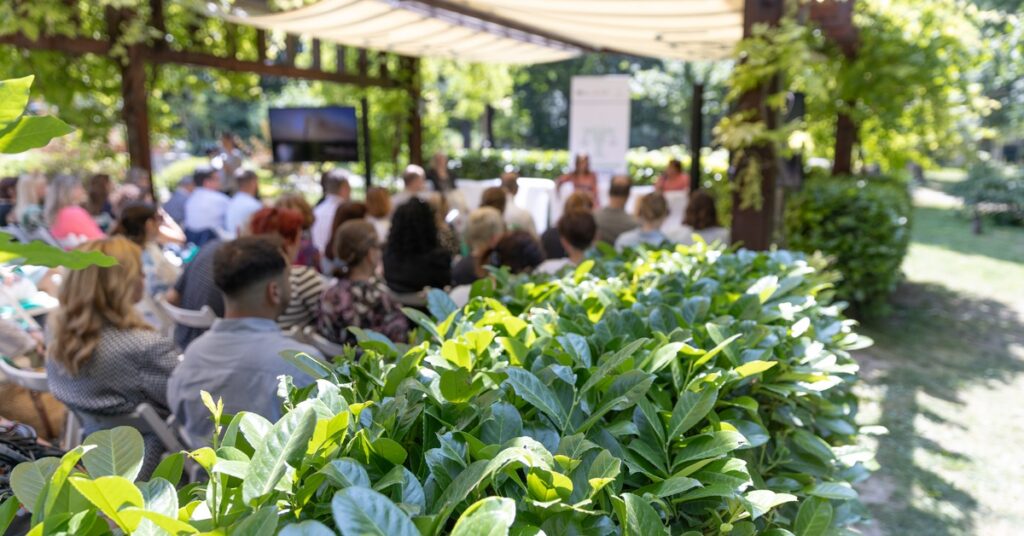 A close-up of the hedges that surround a garden venue with rows of seated guests listening to a presentation.