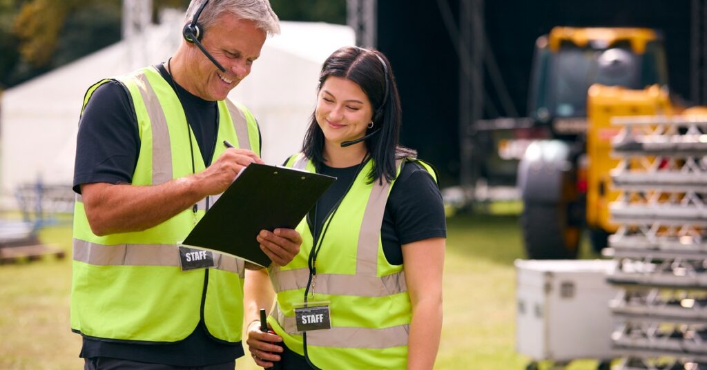 A man and a woman in high-visibility vests, wearing headsets and lanyards that say "STAFF," smile at a clipboard.