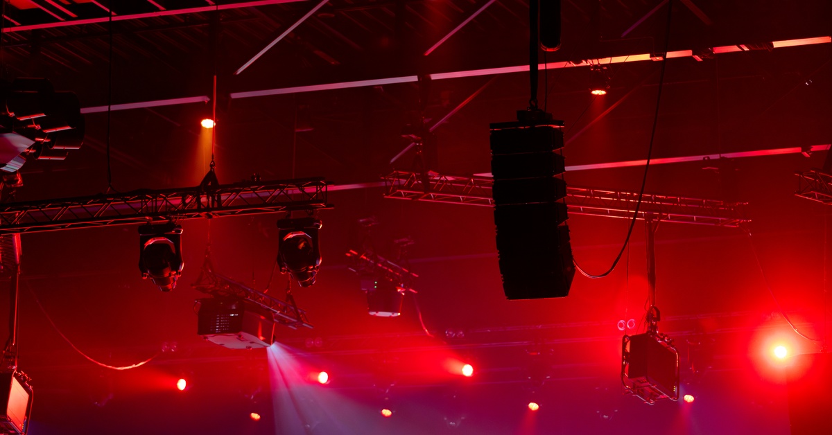 A close-up of the ceiling of a venue production with stage lighting and sound equipment bathed in red light.