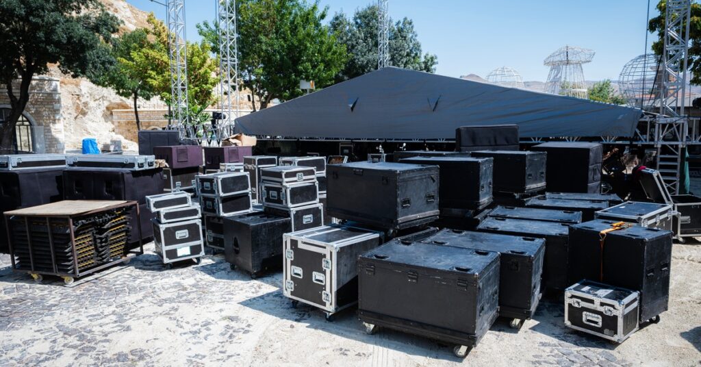 A view behind the stage of an outdoor festival production with flight cases and sound equipment underneath trusses.