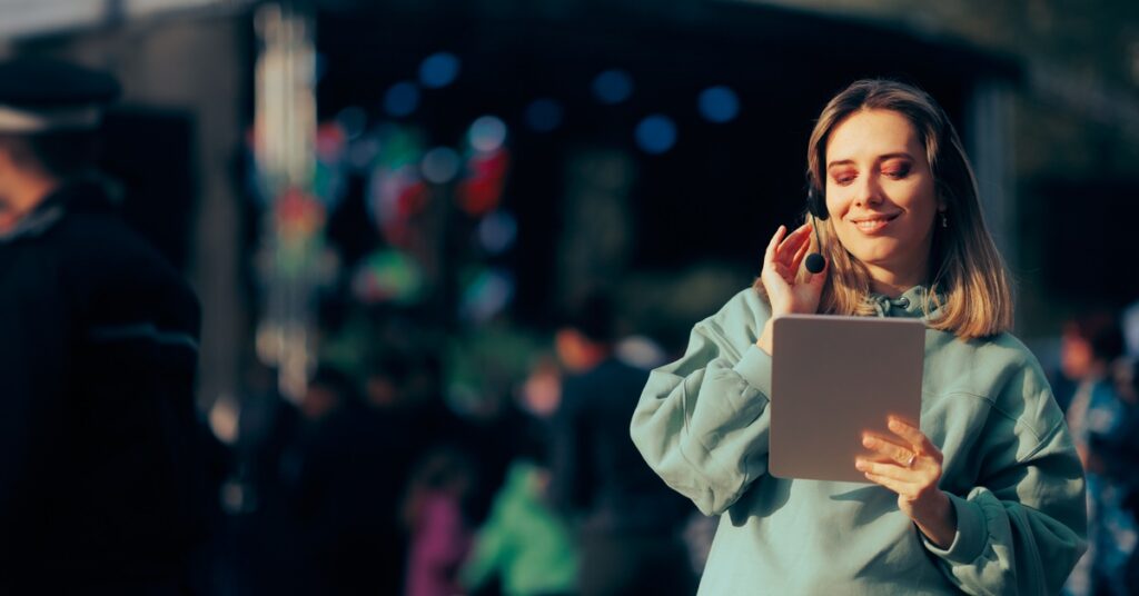 A woman with short blonde hair and a hoodie touches her headset while holding a PC tablet in front of an outdoor stage.